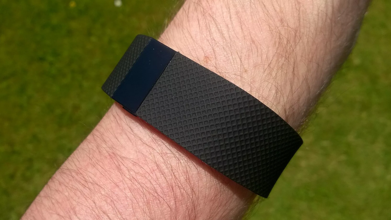 Fekete Fitbit Charge HR aktivitásmérő egy férfi csuklóján, zöld fű előtt