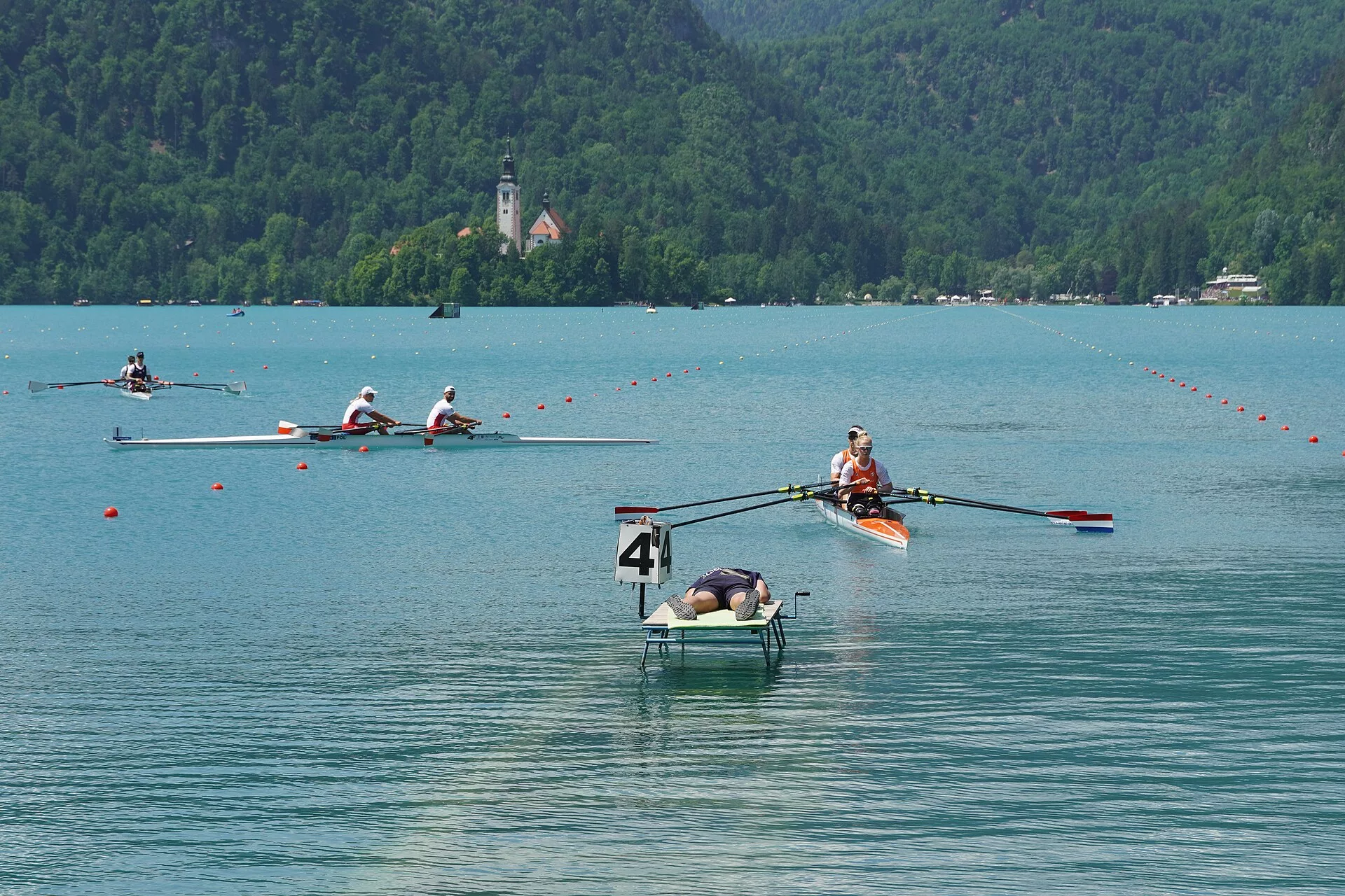 Veterán evezősök hajói a Bledi-tavon egy masters regattán, háttérben a hegyekkel